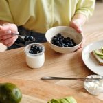 A person prepares a healthy breakfast with yogurt, blueberries, and avocado toast.